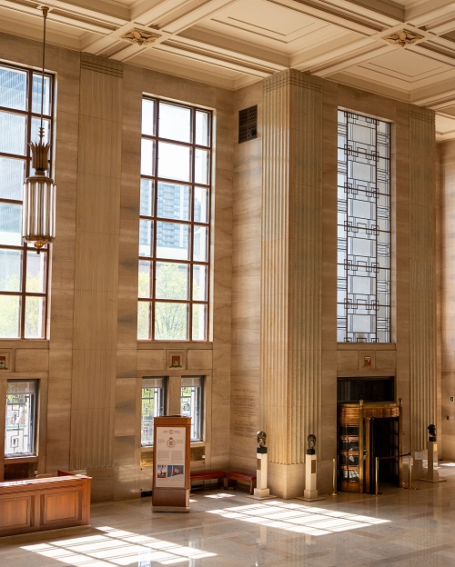 Photo des fenêtres du grand hall d’entrée et d’un luminaire, comprenant un affichage informatif et trois bustes au sol.